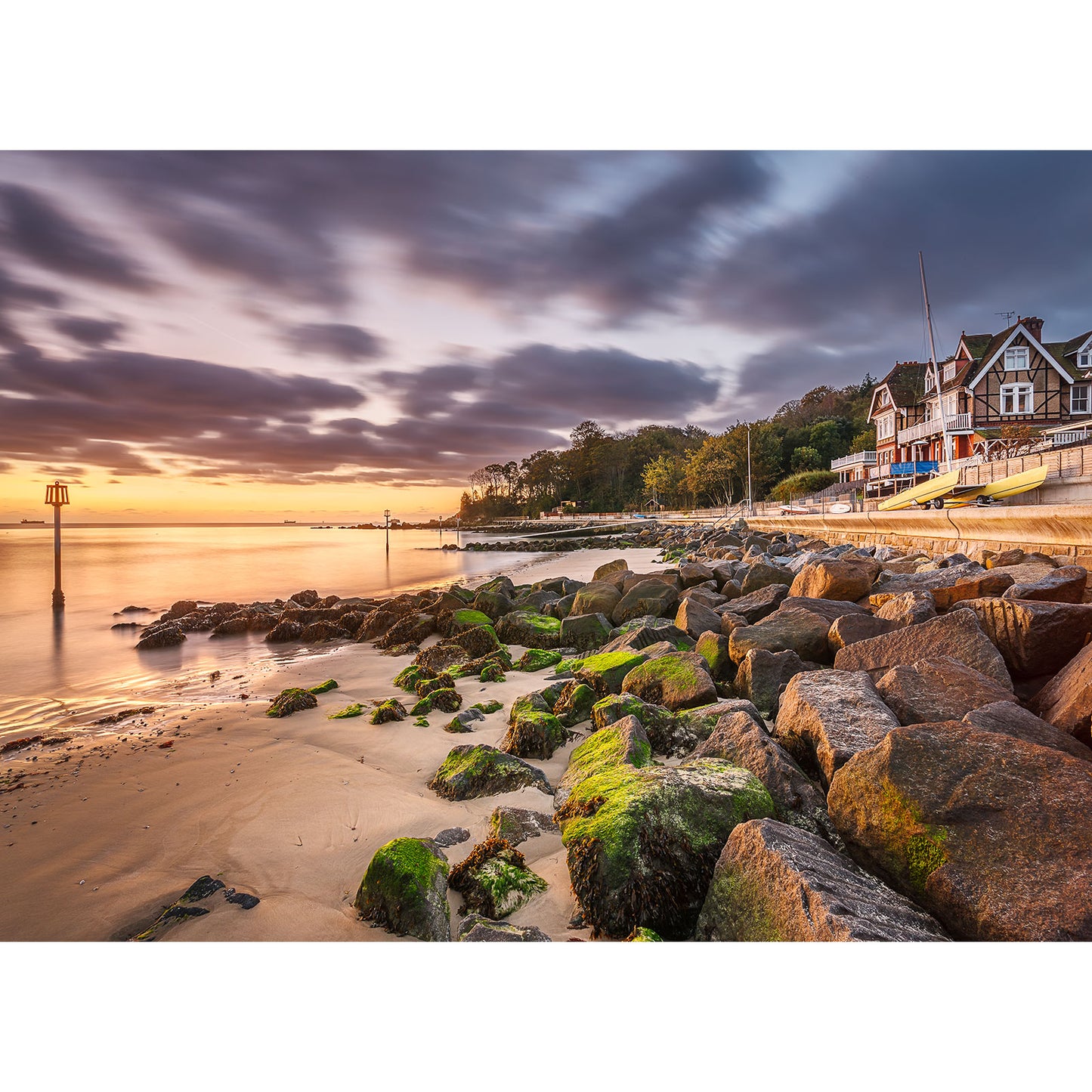 As the late sunrise paints the sky, Seagrove Bay glows with a warm hue, its rocky shoreline framed by houses and a tree-lined path, captured by Available Light Photography.