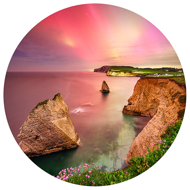 The Northern Lights Freshwater bauble by Available Light Photography features a circular image of a coastal landscape with tall rock formations rising from serene waters, a grassy cliffside adorned with pink flowers, and a vibrant sky shifting from orange to purple.