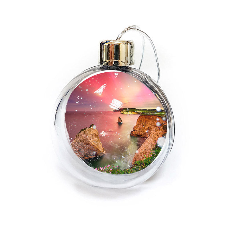 A Northern Lights Freshwater bauble by Available Light Photography featuring a scene of a sunset over rocky cliffs by the sea, complete with snowflakes inside.