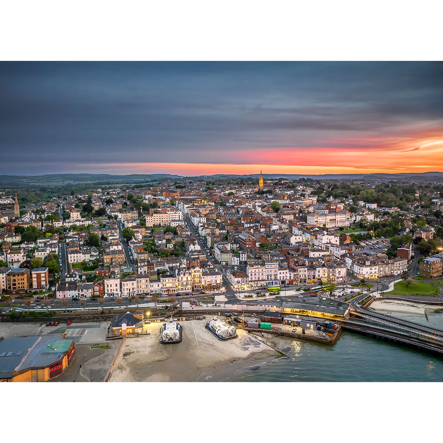 Aerial view of a coastal city at dusk, Ryde by Available Light Photography, featuring a mix of residential and commercial buildings, a waterfront area with sandy patches, and a calm sea under a cloudy sky.