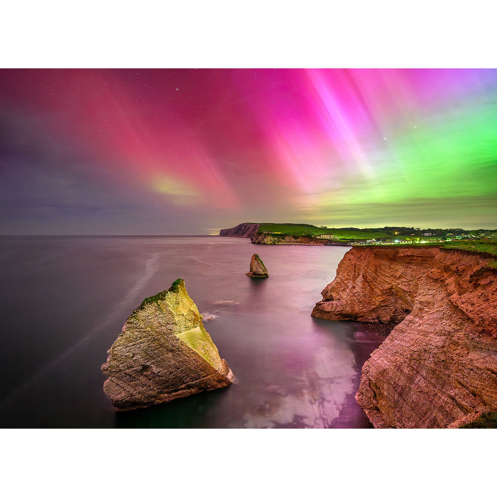 Experience the stunning coastal cliffside of the Isle of Wight, where large rock formations rise from the ocean under vibrant, colorful aurora borealis lights in "The Northern Lights, Freshwater Bay" by Available Light Photography.