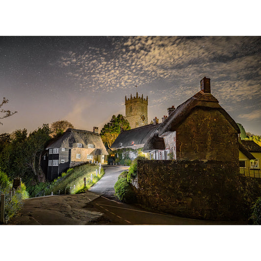 A nighttime photograph from 1955 captures a quaint village with illuminated cottages and Godshill Church in the background, under a partly cloudy sky with visible stars, by Available Light Photography.