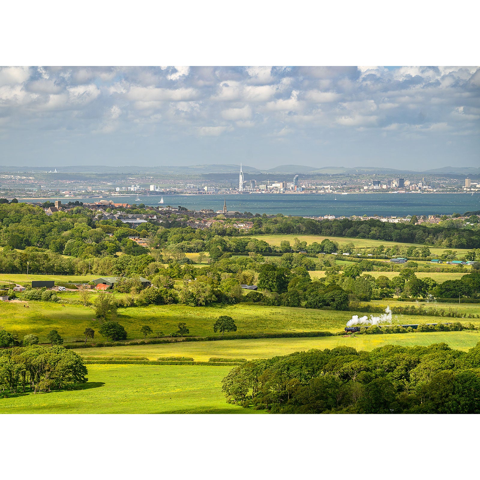 A landscape photo, image number 2700, showing green fields and trees in the foreground, with a city and a water body in the background under a cloudy sky from **View from Ashey Down** by **Available Light Photography**.