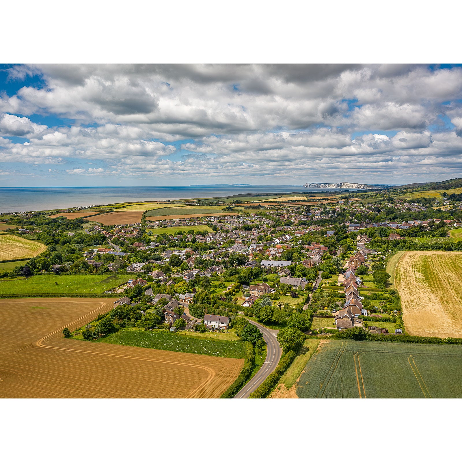 Aerial view of a small village surrounded by fields, with a coastal line and distant hills under a partly cloudy sky. This Brighstone by Available Light Photography description corresponds to image number 1494.