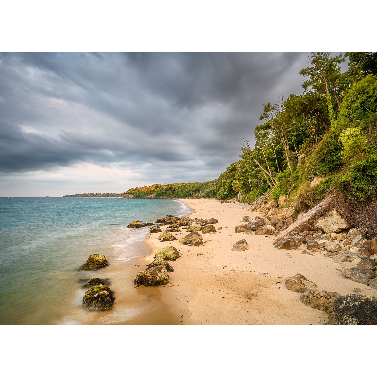 A sandy beach lined with rocks and bordered by dense greenery, under a cloudy sky with an overcast atmosphere. Waves gently lap against the shore, creating an image reminiscent of tranquility like Priory Bay by Available Light Photography.