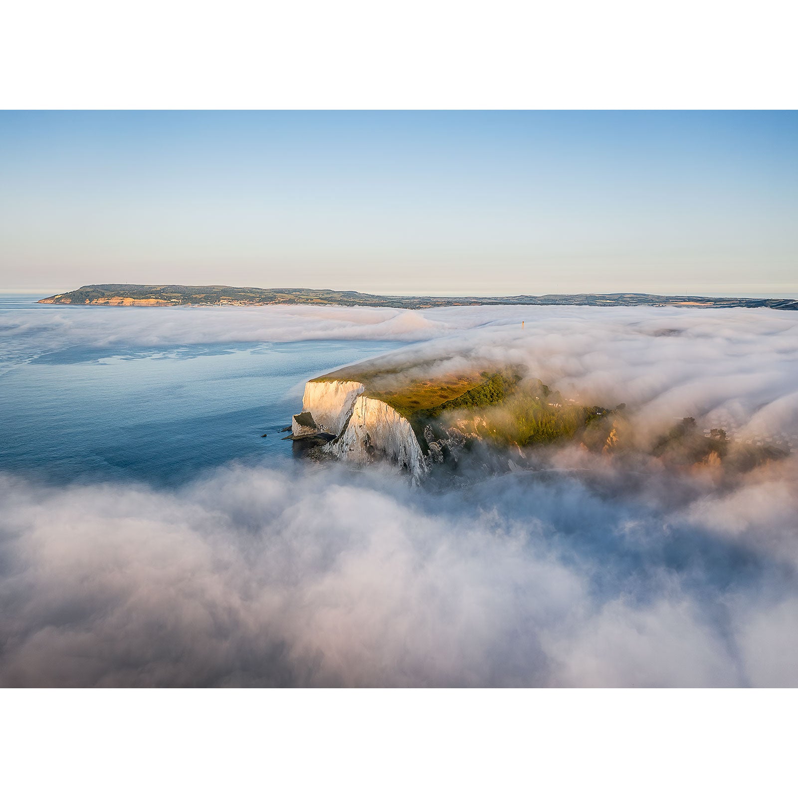 An aerial view of a coastal cliff surrounded by low-lying clouds with the sea and distant landmass in the background under a clear sky. Morning Fog, Culver Cliff by Available Light Photography captures this breathtaking scenery perfectly.