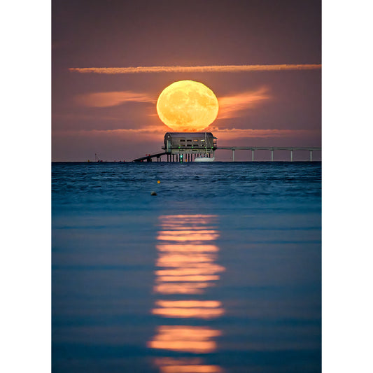 A full moon rises behind a house on a pier over the ocean, casting a bright reflection on the water's surface, reminiscent of Moonrise, Bembridge Lifeboat Station by Available Light Photography.