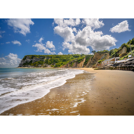 A sandy beach with gentle waves, white cliffs, and greenery in the background under a partly cloudy sky dotted with fluffy clouds. Several structures are visible near the cliffs at Whitecliff Bay by Available Light Photography.