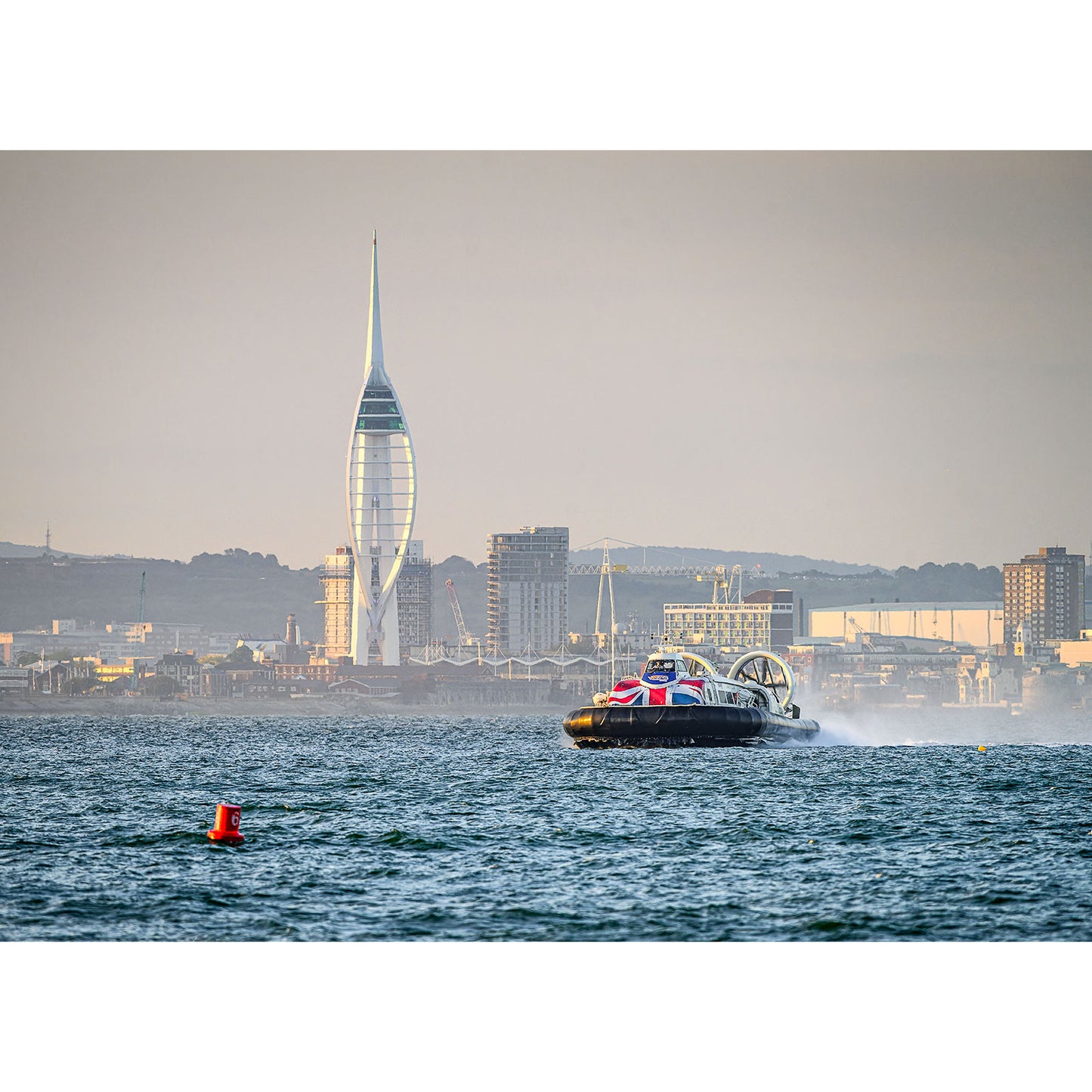 A hovercraft, the Incoming Hovercraft, Ryde by Available Light Photography, moves across the water with a cityscape and the distinctive sail-shaped Spinnaker Tower visible in the background, making for an ideal image number to feature in your product description or metadata.