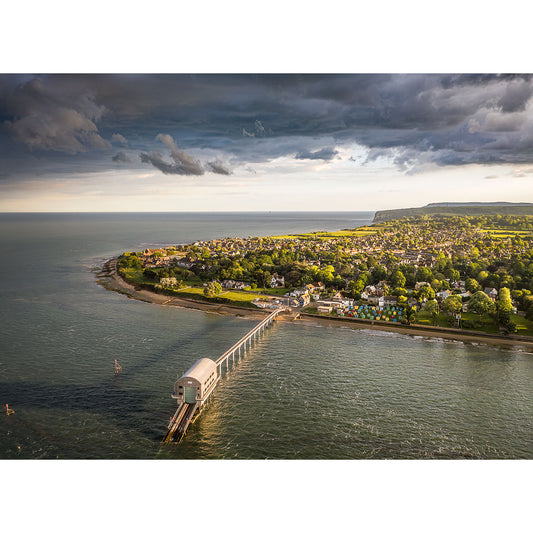 Aerial view of a coastal town with a pier extending into the sea, connected to Bembridge Lifeboat Station by Available Light Photography at the end. Green fields and houses are visible under a partly cloudy sky, enhanced by glorious shades of a stunning sunset.