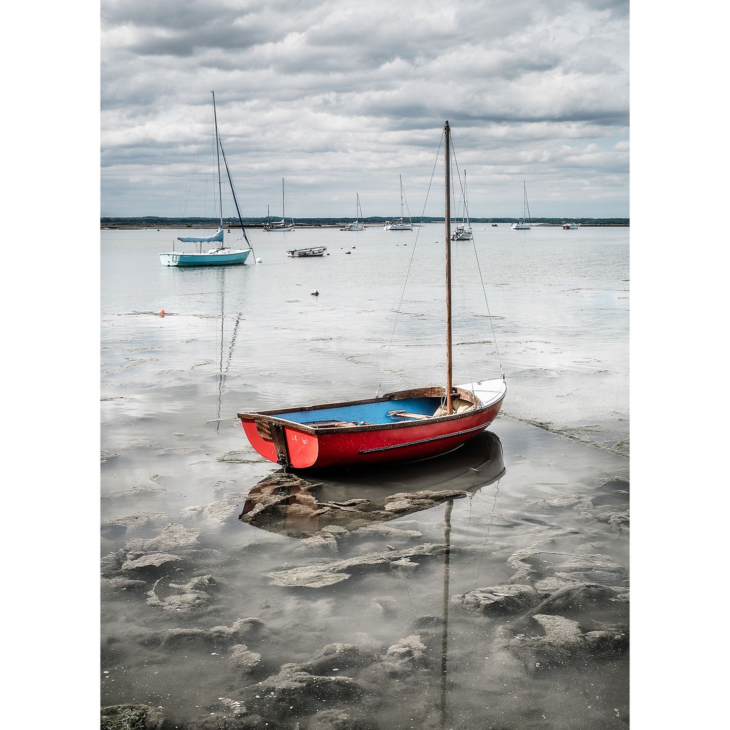 A red and white sailboat is docked in a shallow, muddy area of a body of water with several other boats anchored in the background under a cloudy sky, reminiscent of Available Light Photography's Newtown Creek.