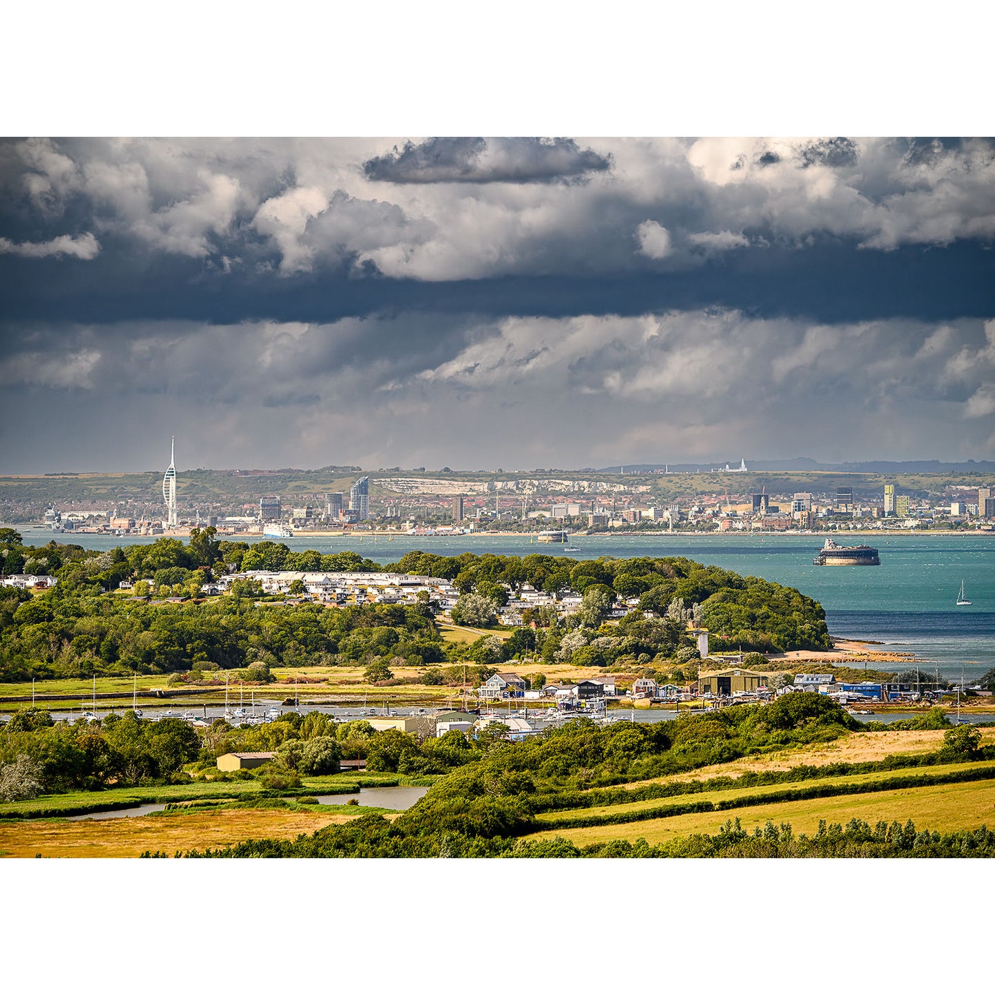 Coastal landscape with a town and harbor in the background, surrounded by greenery under a dramatic, cloudy sky, overlooking Whitecliff Bay and the striking Culver Cliff. This is "View over East Wight" by Available Light Photography.