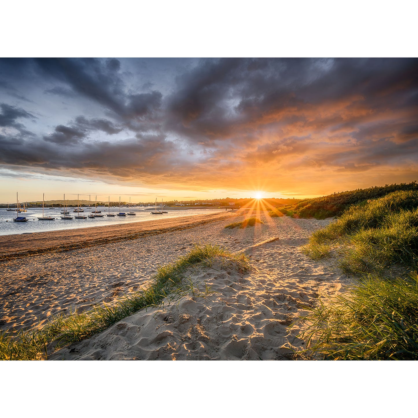 Bembridge's sandy beach features grass-covered dunes at sunset. The sky is dramatic with dark clouds and vibrant orange hues, while boats drift in the water. The sun, partly hidden by the horizon, creates a scene worthy of an Available Light Photography Bembridge Harbour print.