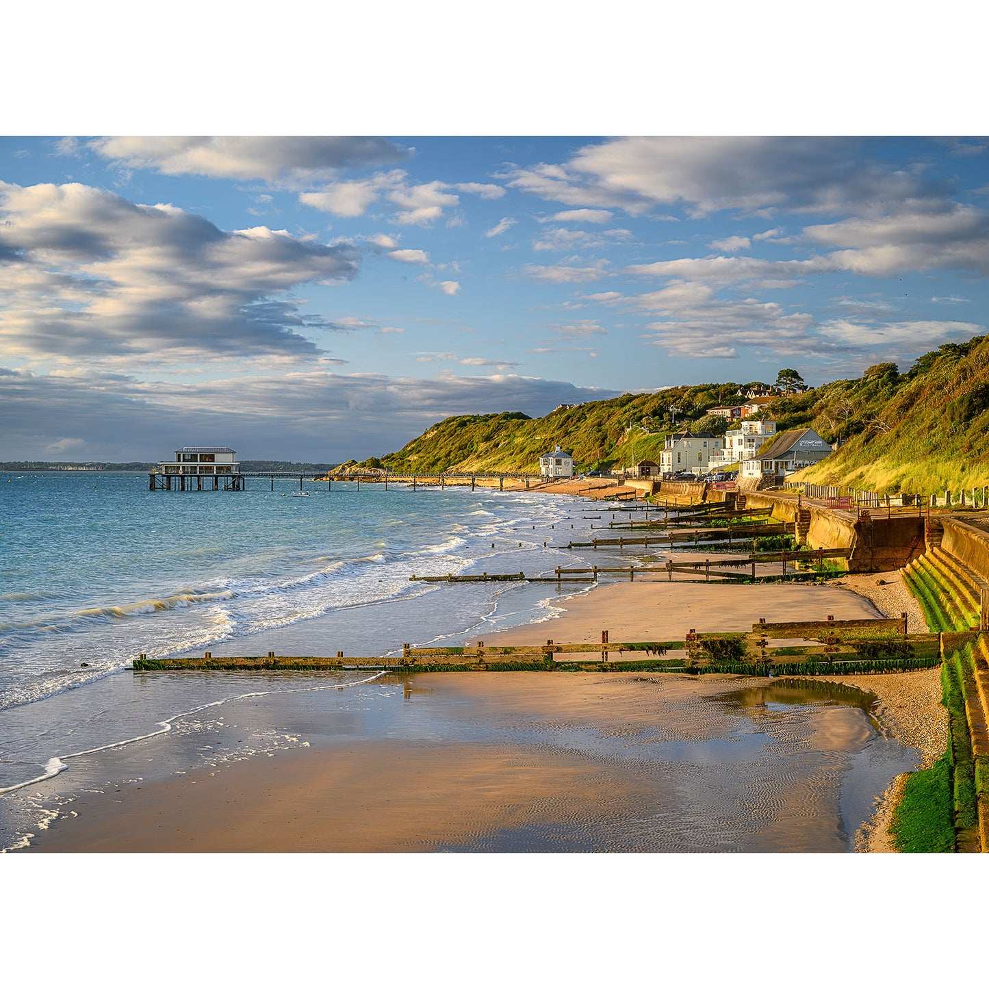 Image number 2709 captures a coastal landscape with a sandy beach, gentle waves lapping against wooden groynes, charming houses on a green hillside, and a pier extending into the sea under a partly cloudy sky. Totland Bay by Available Light Photography.