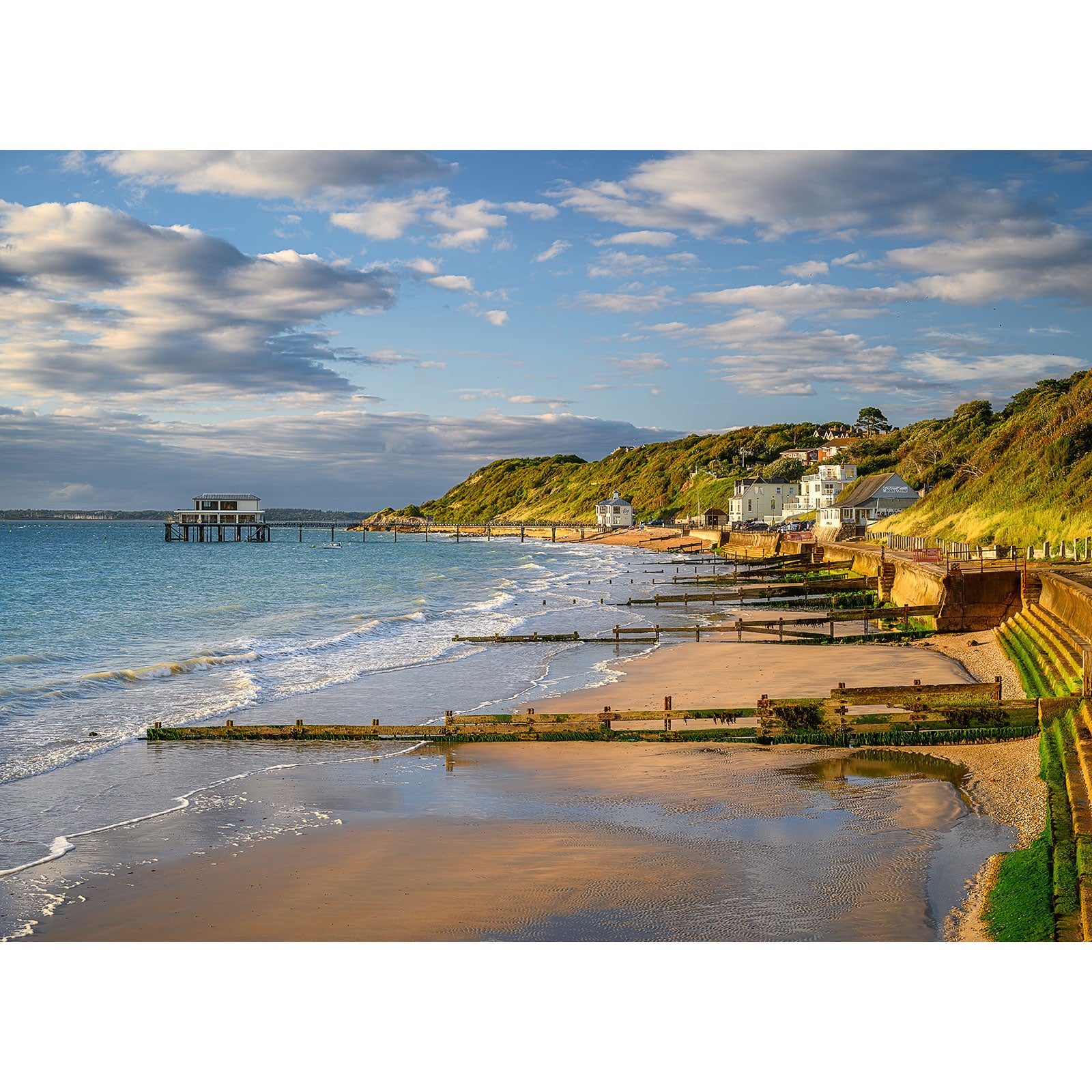 Image number 2709 captures a coastal landscape with a sandy beach, gentle waves lapping against wooden groynes, charming houses on a green hillside, and a pier extending into the sea under a partly cloudy sky. Totland Bay by Available Light Photography.