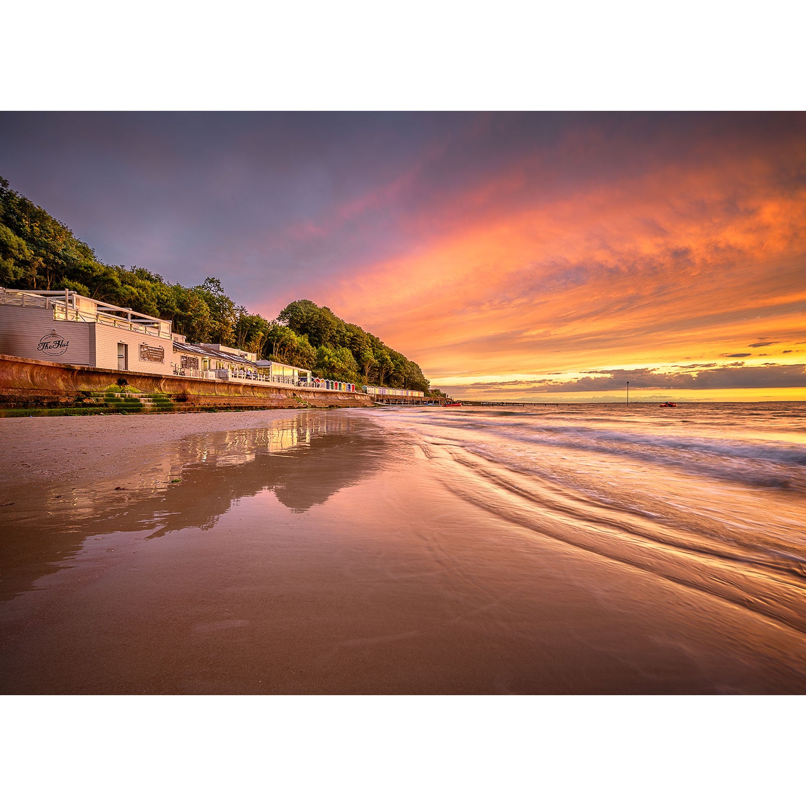 Image number 2892 from Available Light Photography's "Colwell Bay" collection depicts a serene beach scene at sunset. The colorful sky reflects beautifully on the wet sand, with buildings and greenery visible along the shore and gentle waves lapping at the coastline.