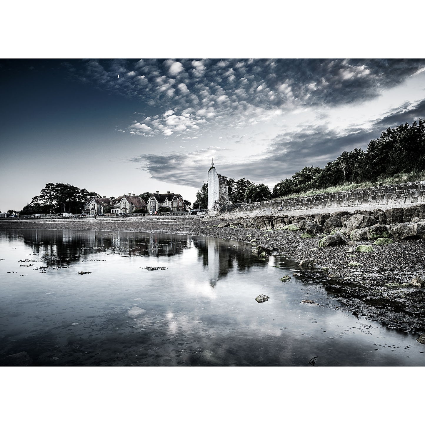 The serene coastal scene of St. Helen's Beach by Available Light Photography captures calm waters reflecting a cloudy sky. Featuring a stone pathway, trees, and buildings under partly cloudy skies, it's the perfect setting for lazy summer days.