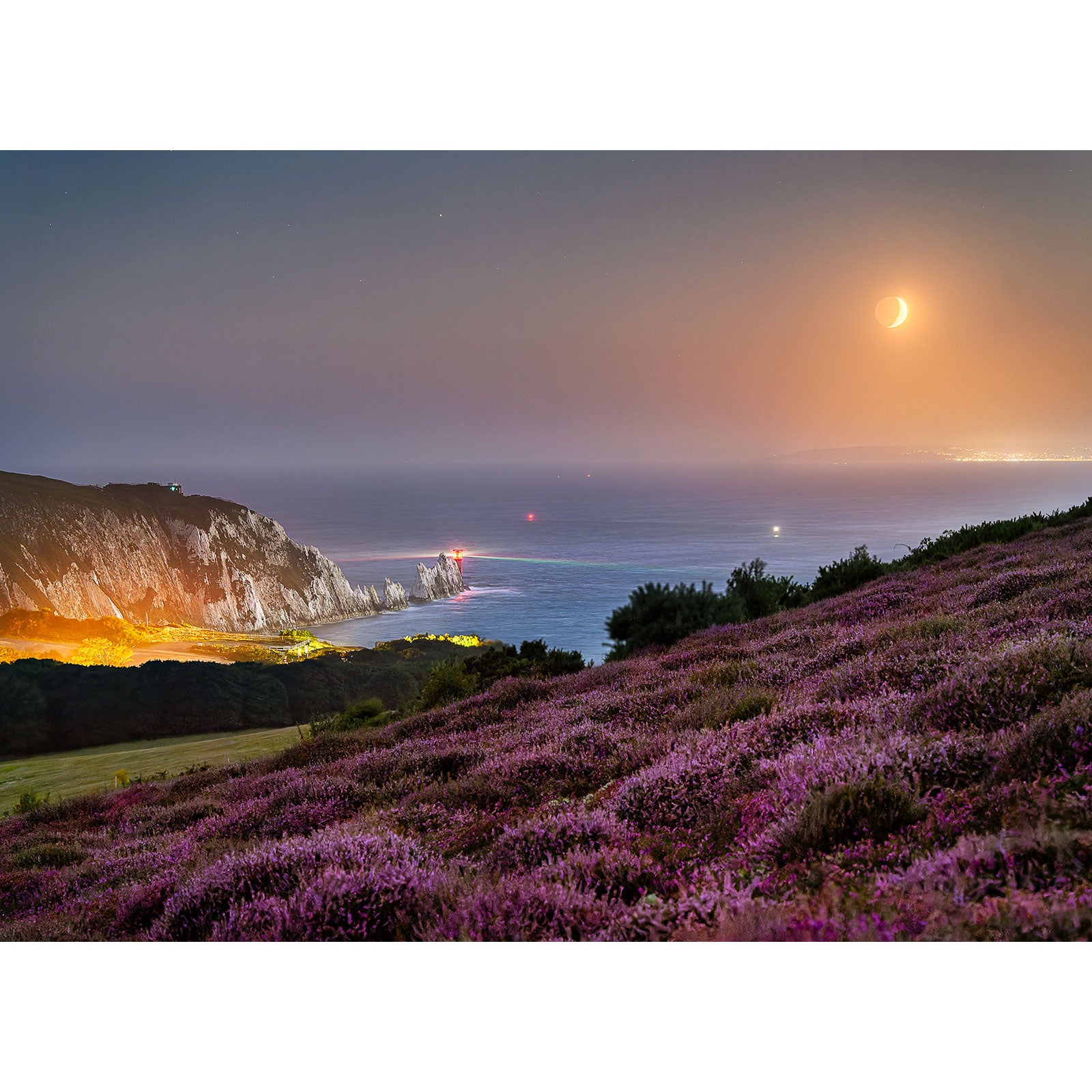 The product "Crescent Moonset over The Needles" by Available Light Photography captures a scene where a crescent moon rises over a coastal cliff adorned with heather in the foreground, with The Needles illuminated by distant lights in the background.