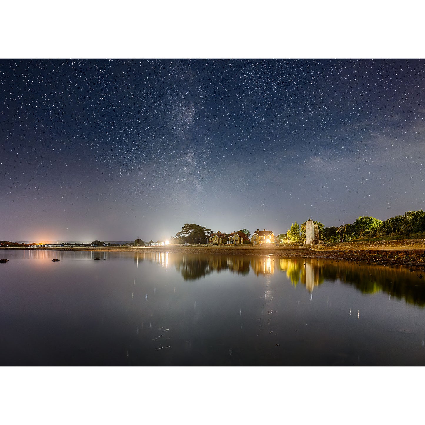 The Milky Way over St. Helen's Beach by Available Light Photography captures a tranquil night at a lakeside village, with a starry sky and lights reflecting on the water, highlighting distant buildings and a peaceful pier.