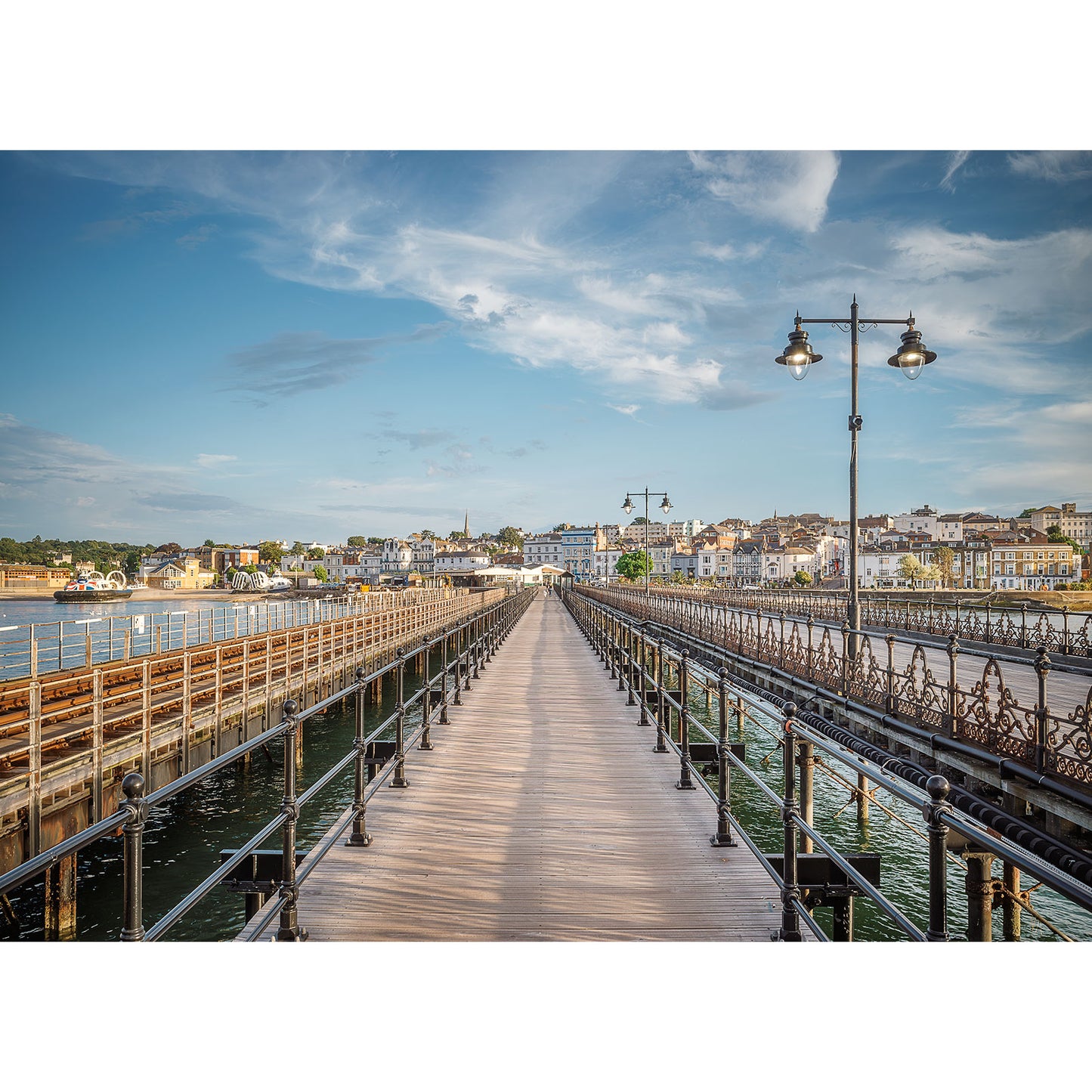 A long, scenic wooden pier—Ryde Pier by Available Light Photography—stretches majestically over the water towards a distant town, with a blue sky adorned with scattered clouds overhead. Two ornate street lamps are strategically placed along the pier, forming an idyllic scene ideal for your next product description or SEO keyword use.