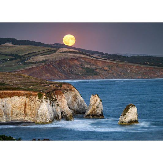 Moonrise, Freshwater Bay by Available Light Photography captures a bright full moon rising over a coastal landscape with rugged cliffs and sea stacks, waves gently crashing against the rocks, and hilly terrain in the background.