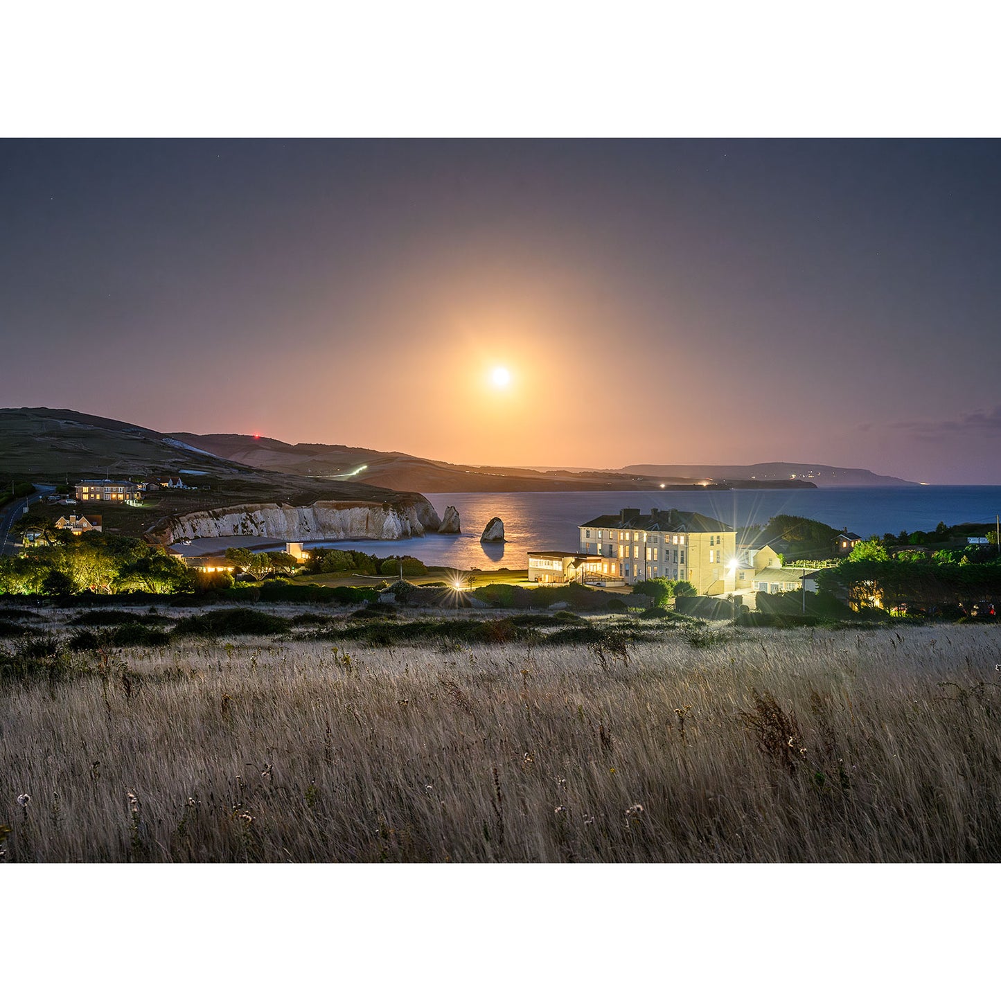 The product named "Moonrise, Freshwater Bay" by Available Light Photography depicts a serene coastal landscape at night, showcasing a bright moon illuminating a cliffside, calm sea, and a lit building by the shore with grassy fields in the foreground. Image number 2905 perfectly captures this tranquil scene.