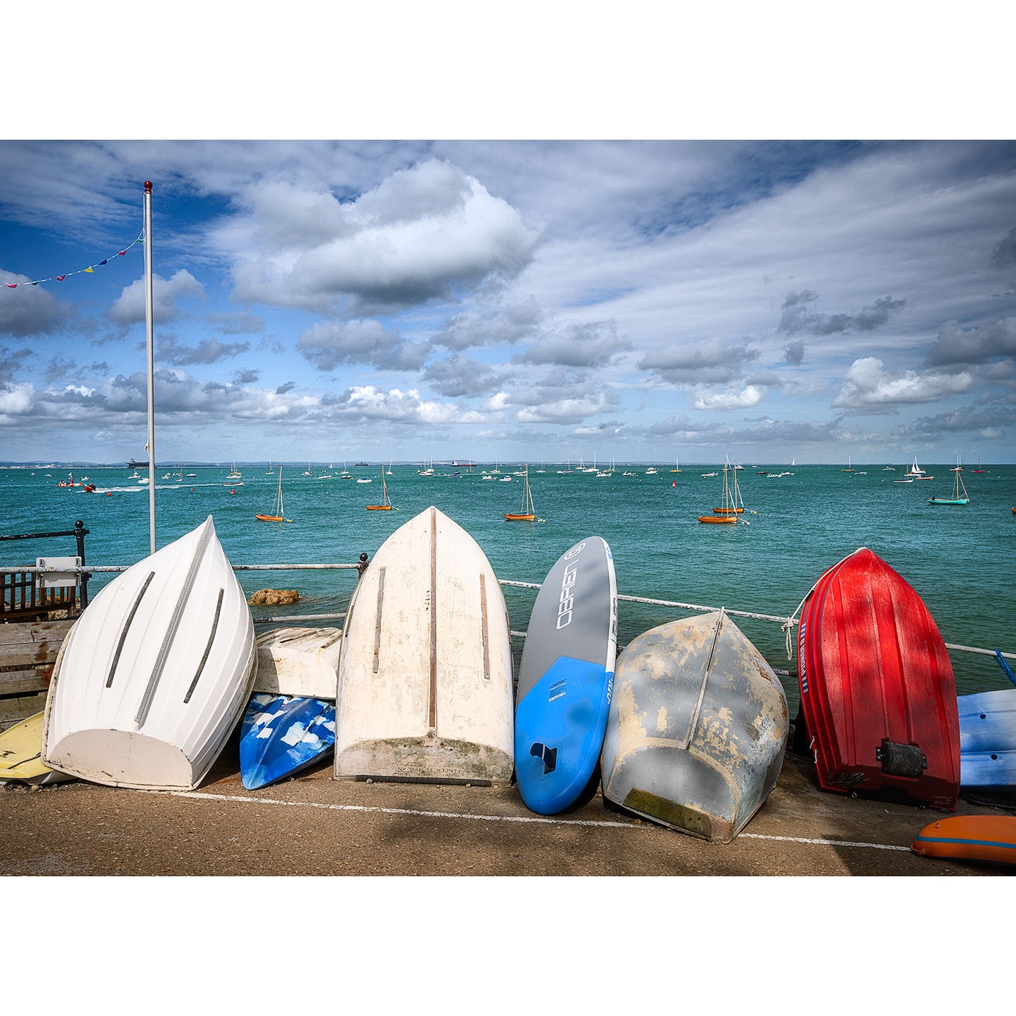 Seaview by Available Light Photography captures a row of upturned small boats resting on a shore with a view of the turquoise sea and sailboats in the distance under a cloudy sky.