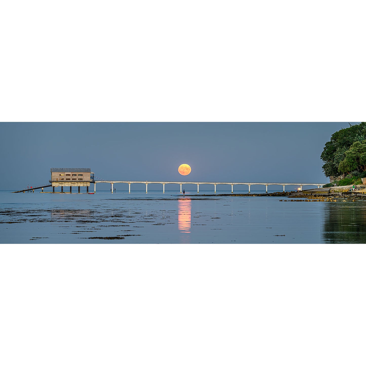 Moonrise, Bembridge Lifeboat Station by Available Light Photography captures a full moon rising over a pier and building extending into a tranquil body of water, with trees visible on the right side. The moon's reflection shimmers on the water's surface, creating an enchanting image reminiscent of serenity.