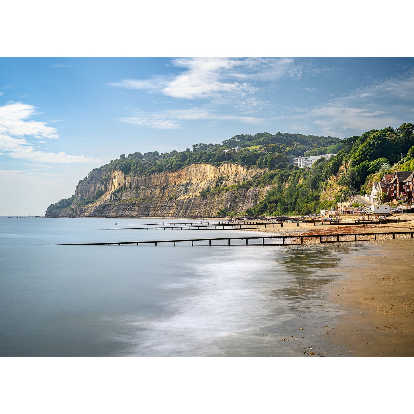 The photograph titled "Shanklin" by Available Light Photography showcases a coastal scene featuring a sandy beach, wooden piers stretching into the sea, and cliffs adorned with lush greenery under a partly cloudy sky.