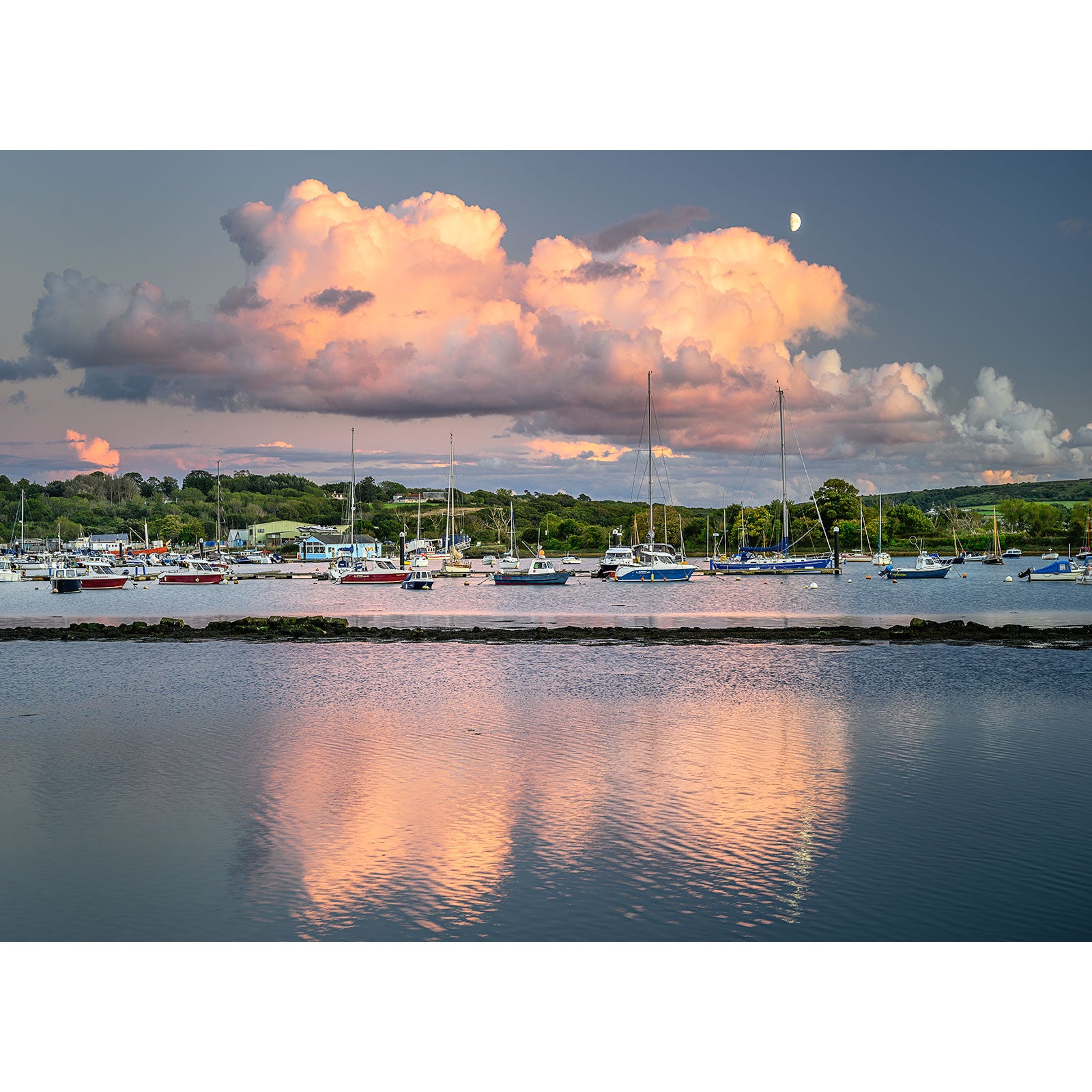 The product titled "Bembridge Harbour" by Available Light Photography showcases a tranquil scene of a harbor filled with numerous sailboats docked on calm water. The photograph captures the beauty of large clouds illuminated by the setting sun, with green hills in the background and serene reflections in the water, creating an idyllic and picturesque setting.