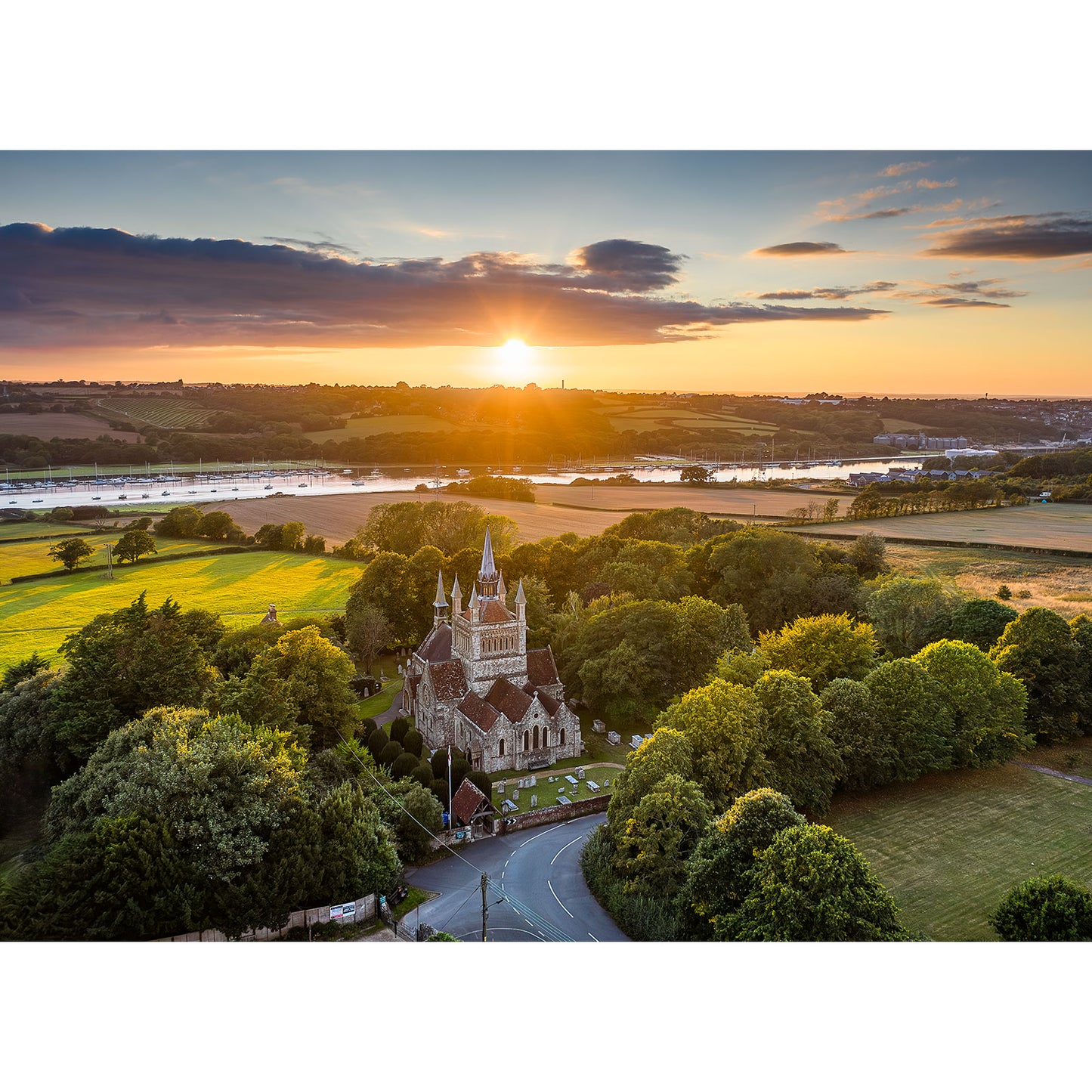 Aerial view of Whippingham Church, surrounded by trees and fields at sunset, with a river and distant countryside in the background. Image number: 2913. Photo by Available Light Photography.