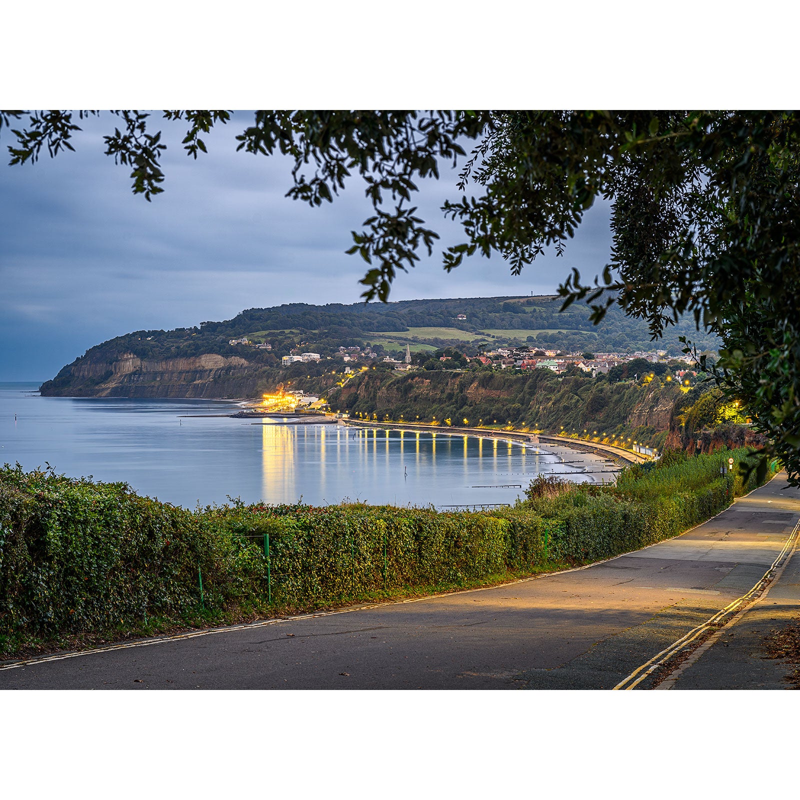 A coastal road with a view of a bay at dusk, featuring illuminated buildings along the shoreline and a hilly landscape in the background, framed by tree branches. This serene image, titled "Looking towards Shanklin" by Available Light Photography, perfectly captures the tranquil beauty of the evening scenery.