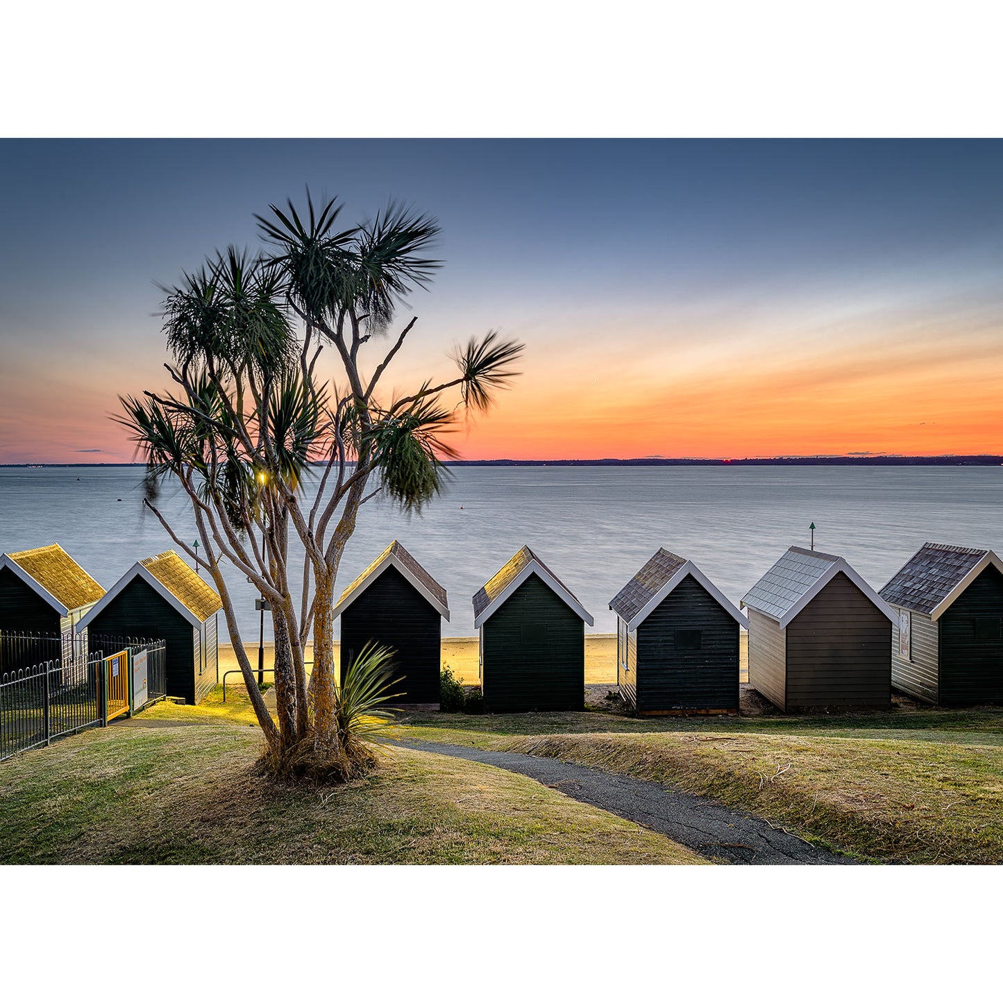 Product Description: The "Gurnard" by Available Light Photography captures vibrant beach huts positioned beside a peaceful sea during sunset, accompanied by an elegant palm tree in the foreground, all under a clear sky that amplifies the soothing atmosphere.