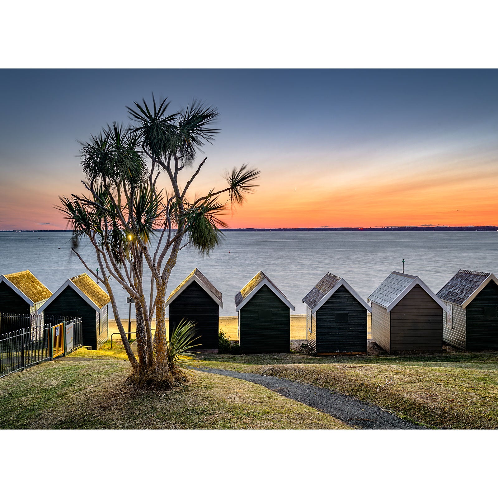 Product Description: The "Gurnard" by Available Light Photography captures vibrant beach huts positioned beside a peaceful sea during sunset, accompanied by an elegant palm tree in the foreground, all under a clear sky that amplifies the soothing atmosphere.