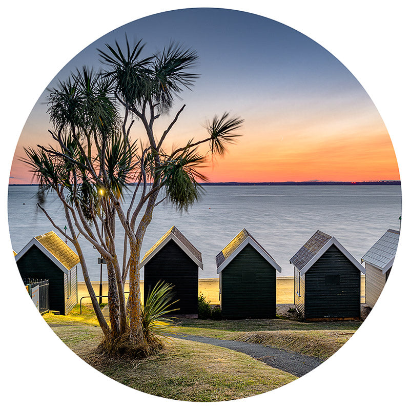 Gurnard bauble by Available Light Photography captures a sunset casting its glow over a row of small, dark beach huts, complemented by a tree in the foreground and calm water stretching into the background.