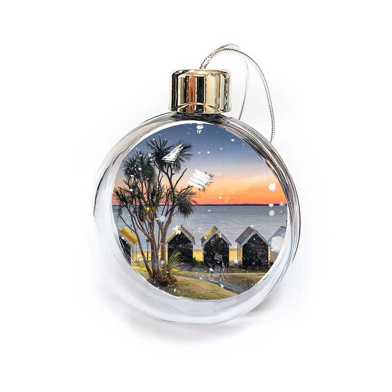 Gurnard bauble by Available Light Photography showcases a coastal winter scene with beach huts, palm trees, a sunset, and snowflakes.