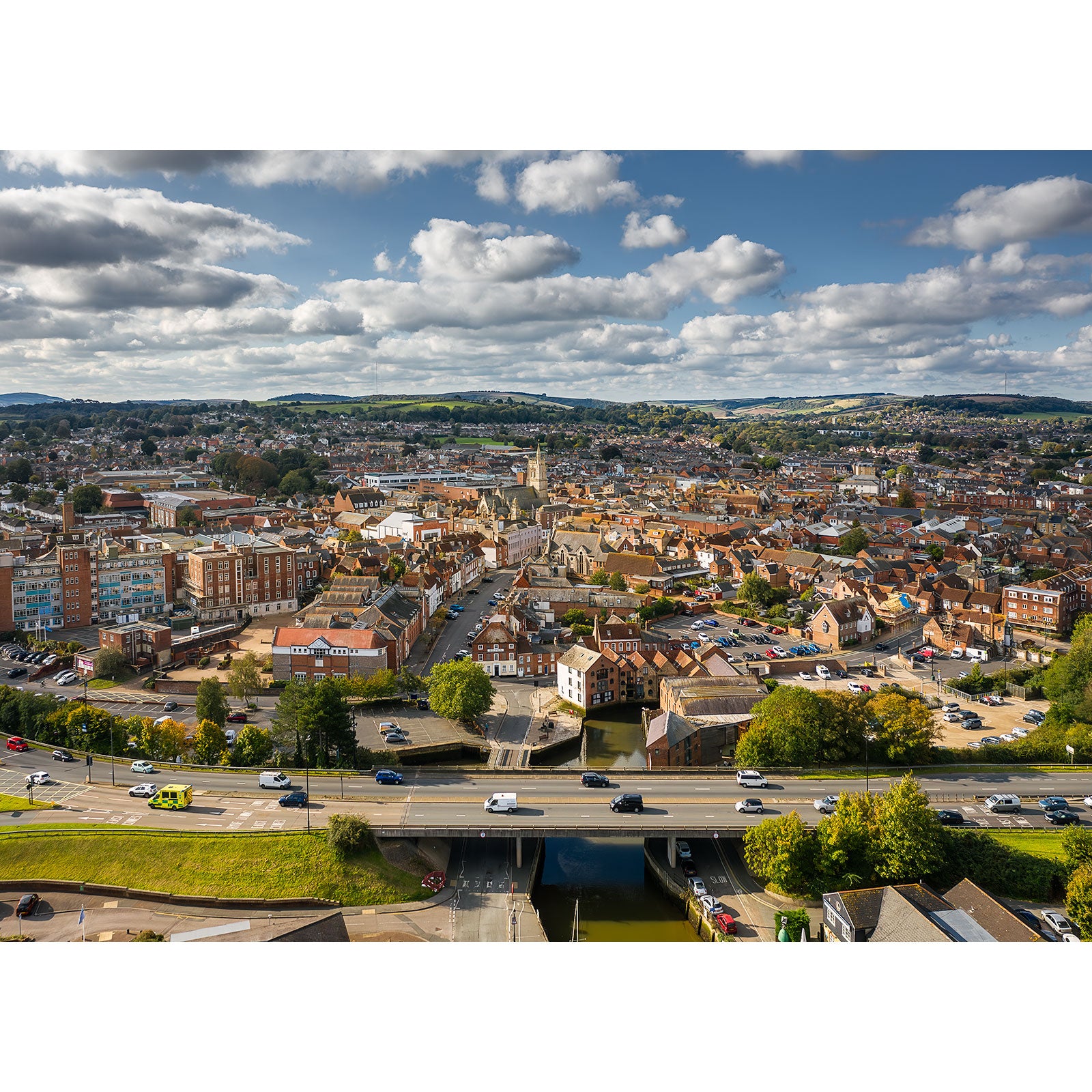 Seen from above, Newport unveils its charm—a blend of historic and modern architecture highlighted by a graceful church steeple, captured beautifully by Available Light Photography. Roads weave through the scene with vehicles moving against lush, verdant hills under a partly cloudy sky, encapsulating an essence of timeless serenity.