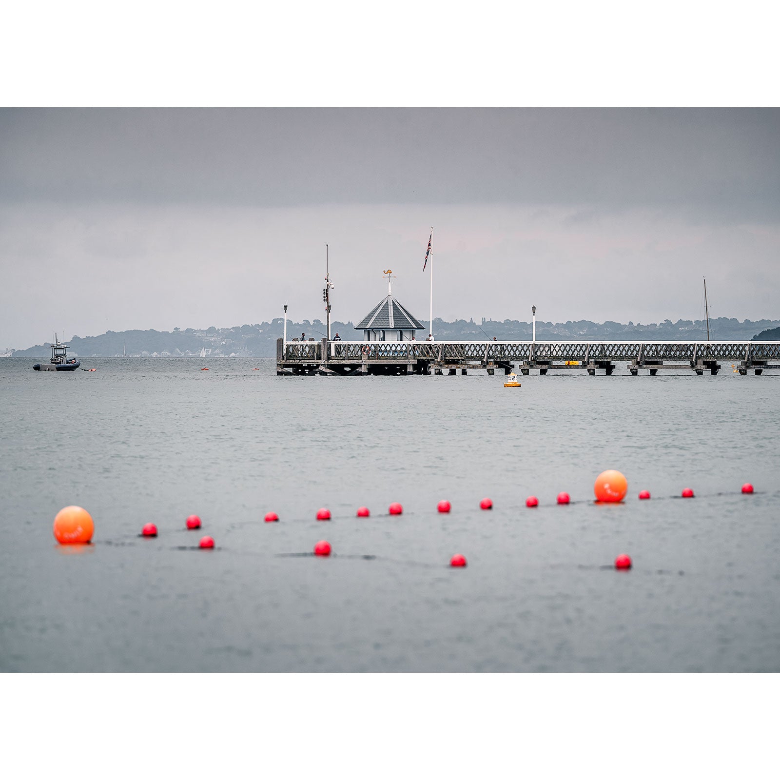 The Yarmouth Pier, captured by Available Light Photography, stretches into a tranquil body of water accented by red buoys in the foreground. To the left, a small boat appears against an overcast sky, infusing the scene with muted serenity.