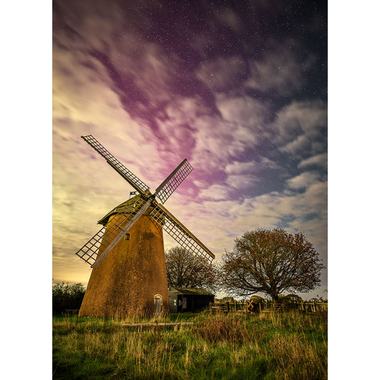 A historic windmill, reminiscent of the enchanting Bembridge Windmill by Available Light Photography, stands majestically under a starry night sky, surrounded by lush grass and trees.