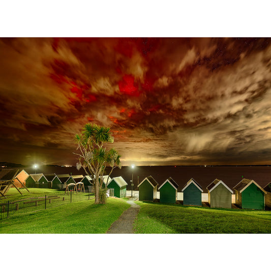 Under a dramatic, cloud-strewn orange night sky, a charming row of huts sits along the shoreline in the "Gurnard" image by Available Light Photography. A winding path weaves through the vibrant green grass with the silhouette of a towering palm tree on the left. (Image number: 2648)