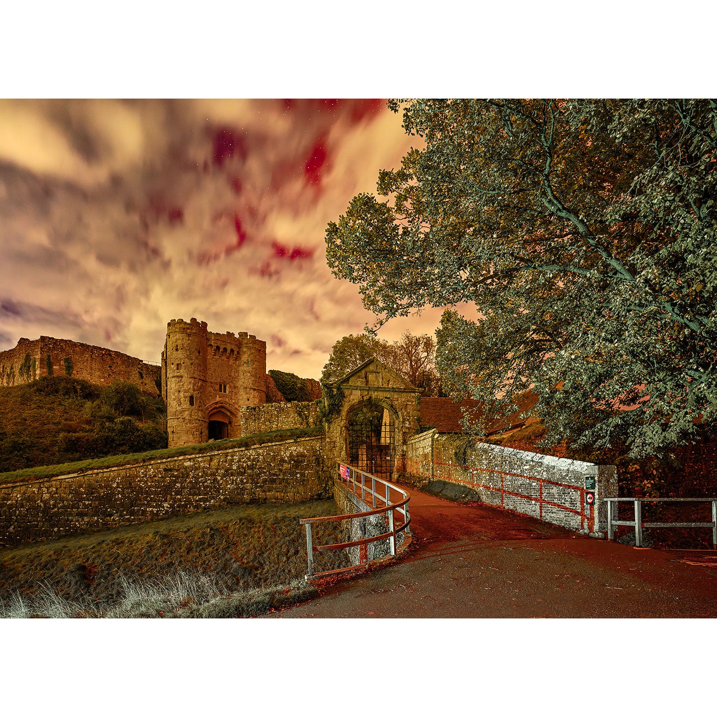 A dramatic sky over an ancient stone castle and bridge, framed by trees, creates an atmospheric scene reminiscent of the Carisbrooke Castle image from Available Light Photography's timeless photo series.