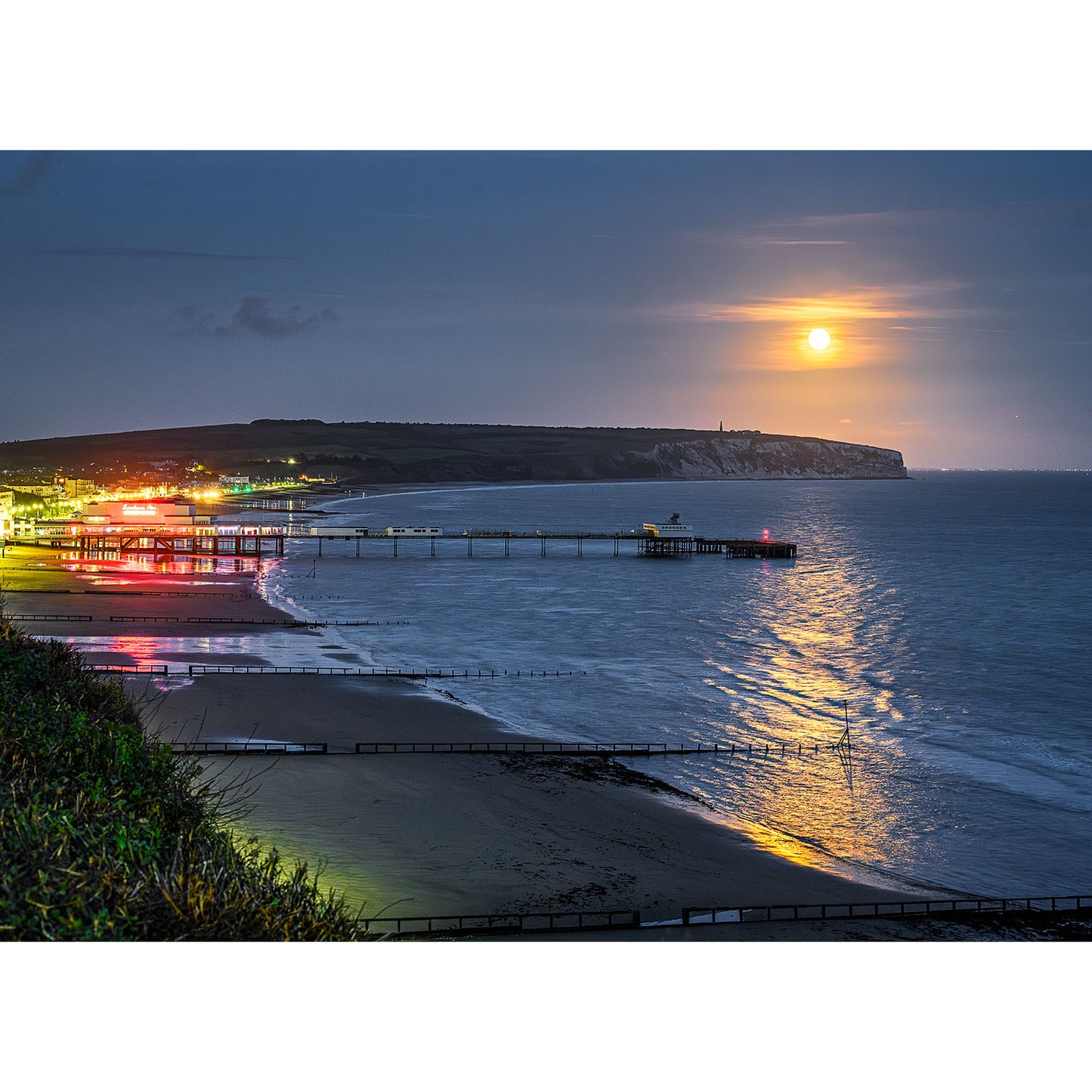 Image number 1784, titled "Moonrise over Sandown Bay" by Available Light Photography, captures a stunning coastal view at night with an illuminated pier and buildings beneath a full moon, their reflections dancing gracefully on the sea.