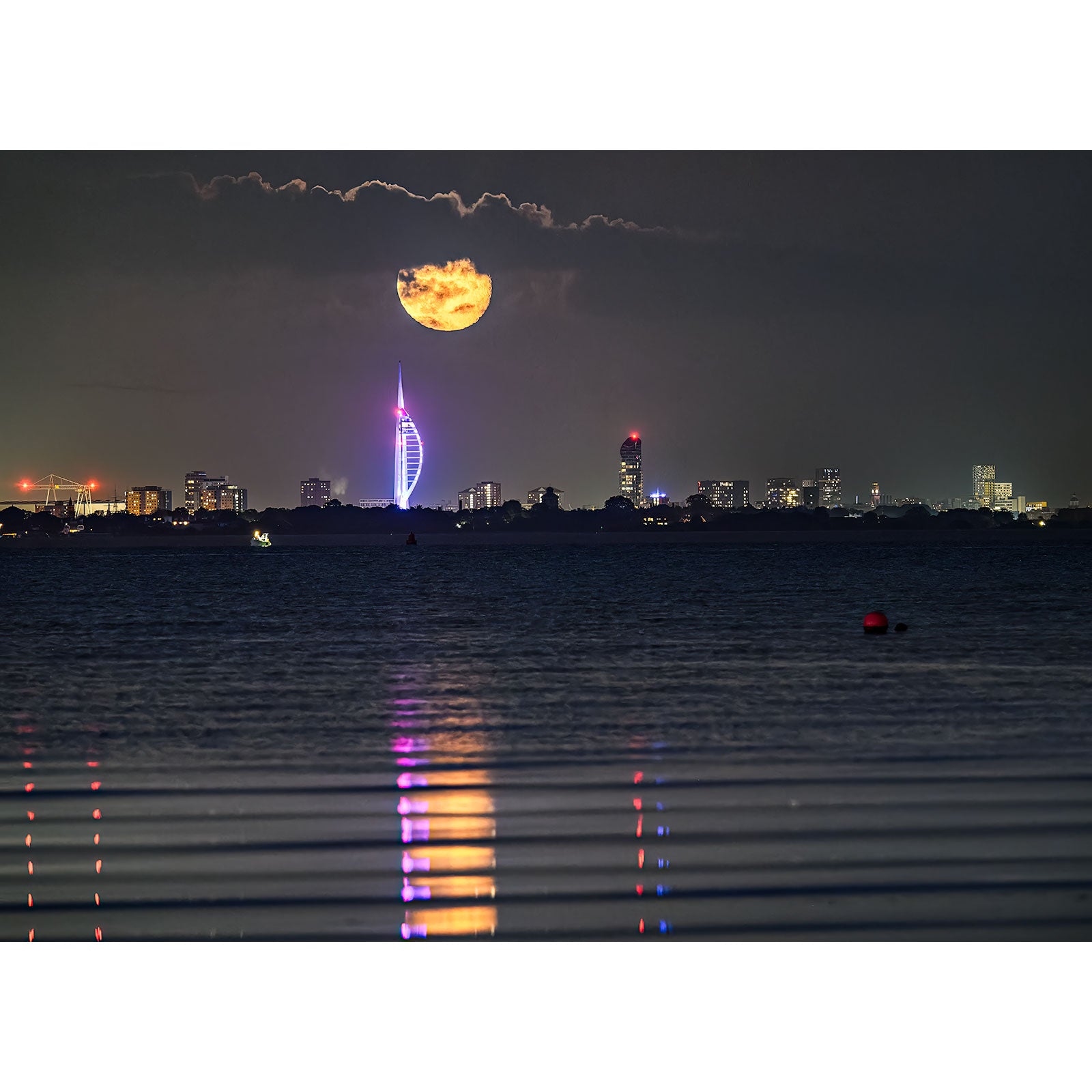 In the photograph "Moonrise, The Spinnaker Tower" by Available Light Photography, a full moon bathes the city skyline in its glow, accentuating the distinct illumination of The Spinnaker Tower. This tranquil scene reflects on the still water below, weaving a magical nocturnal tapestry that stands unapologetically beautiful.