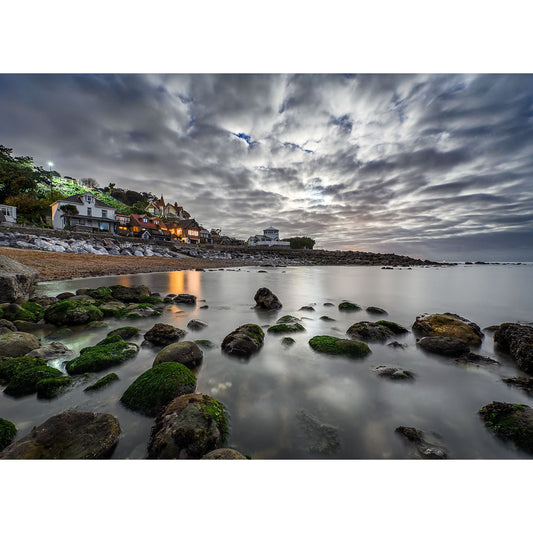 Under a cloudy night sky, "Steephill Cove by Moonlight" from Available Light Photography captures the coastal scene coming alive with a rocky shoreline and seaweed-covered stones, while illuminated houses cast a warm glow over the enigmatic landscape.