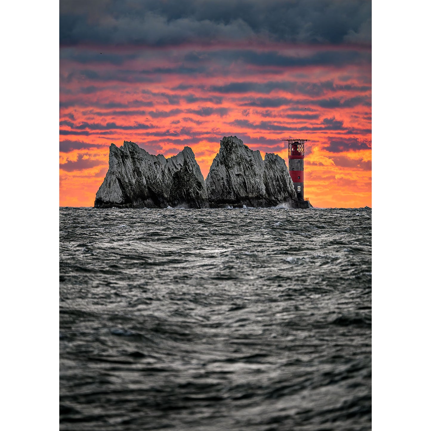 Amidst a rough sea with jagged rocks, The Needles by Available Light Photography captures a solitary lighthouse standing resiliently against the dramatic clouds at sunset, casting its beam over a vibrant orange-red sky.