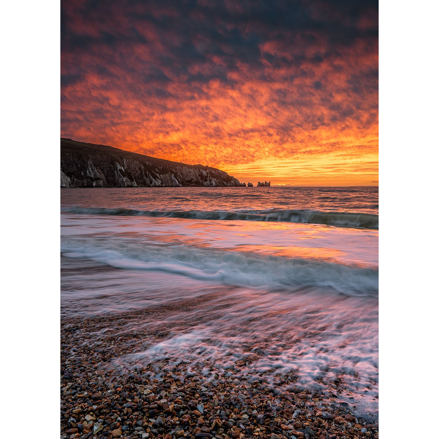 A mesmerizing sunset unfolds over The Needles' rocky shoreline, with waves gently lapping the beach under a dramatic orange and purple sky, creating an enchanting coastal symphony by Available Light Photography.