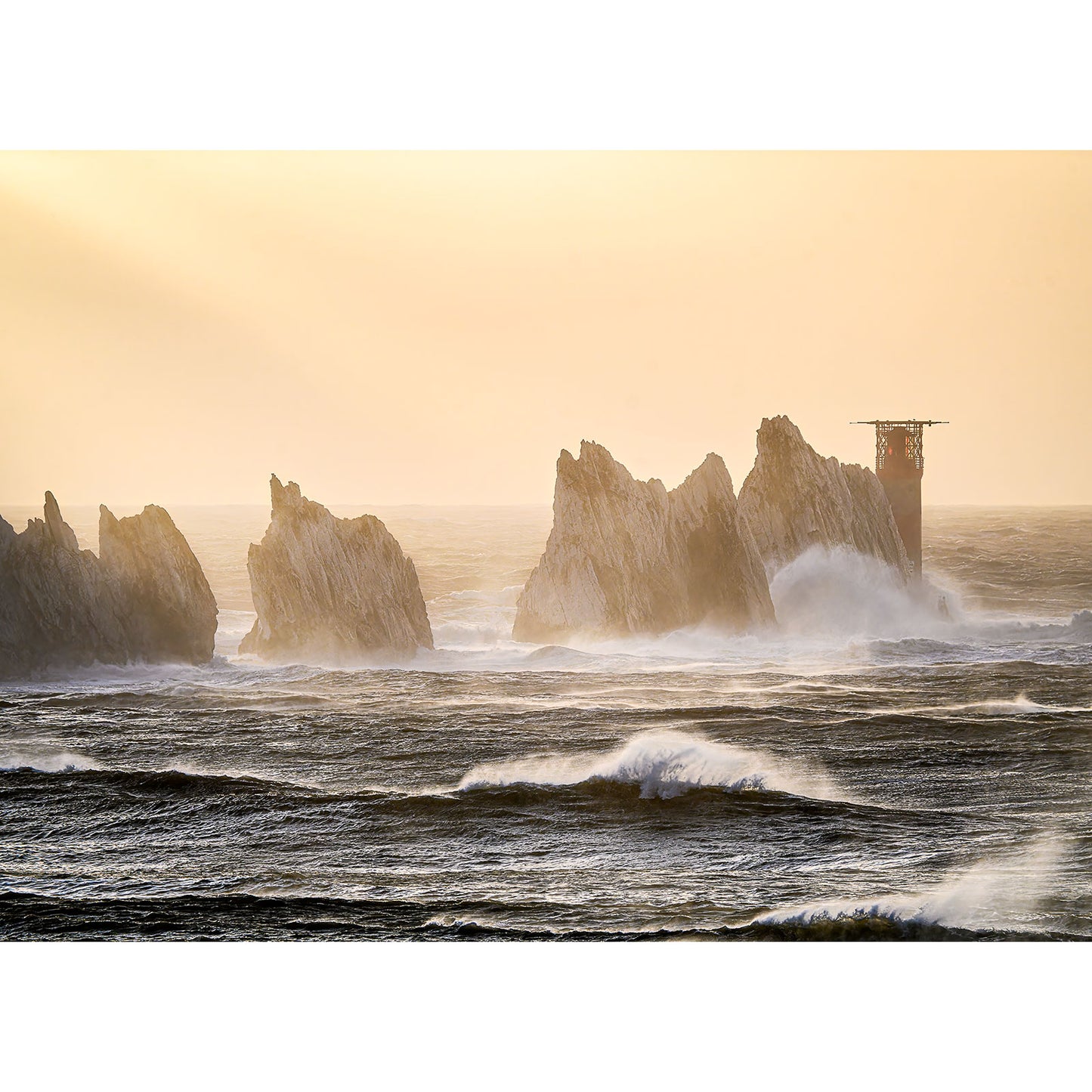 Under a hazy, golden sky, jagged rock formations known as The Needles rise from the sea, waves crashing against them and silhouetted by an orange glow—captured beautifully by Available Light Photography.