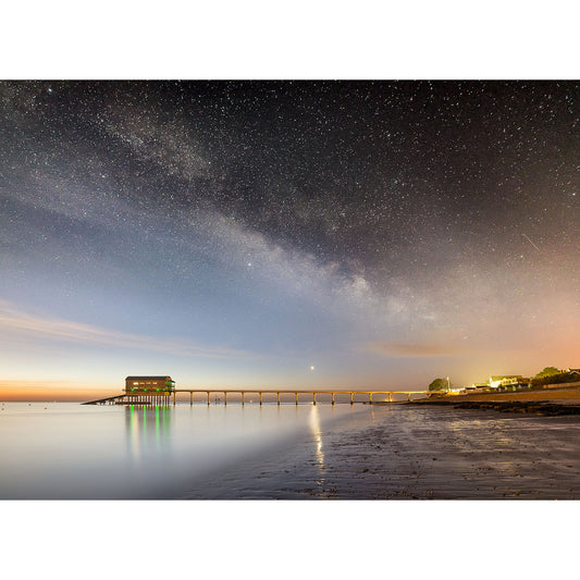 A Milky Way night sky over the serene Isle of Wight beach with a long pier illuminated by lights reflecting on the water's surface captured by Available Light Photography at Bembridge Lifeboat Station.