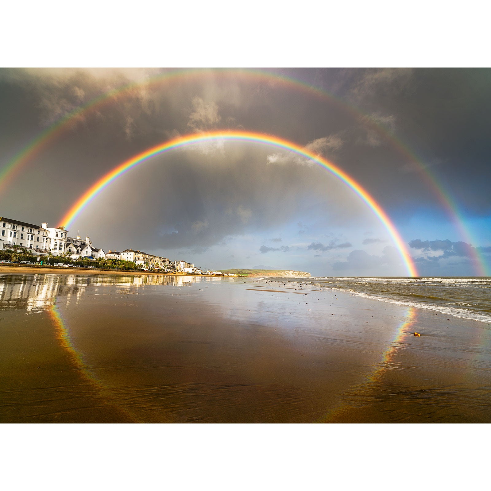 A vivid double Rainbow over Sandown Bay arcs over a beachfront with reflections on the wet sand, as captured by Steve from Available Light Photography.