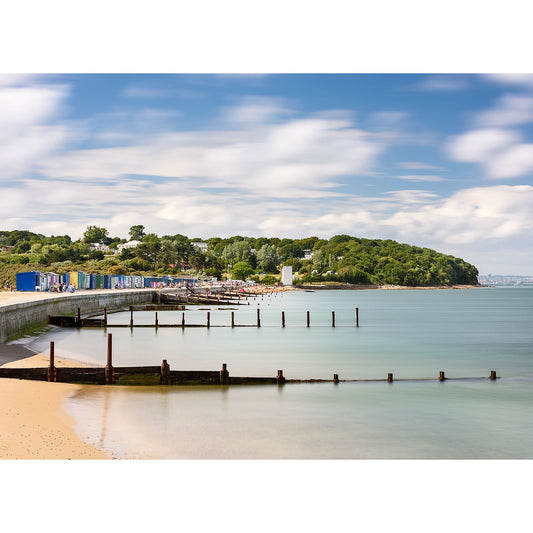 A serene beach scene captured by Available Light Photography's "St. Helen's Beach" embraces the essence of summer with calm waters, a sandy shore, wooden groynes, colorful beach huts, and a tree-covered hill in the background under a partly cloudy sky—perfect for lazy days.